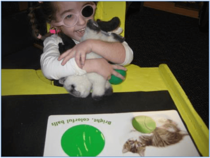 A young girl holds a stuffed kitten with an open book about a kitten on the desk in front of her.
