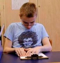 A boy with glasses reading a page of braille text at his desk