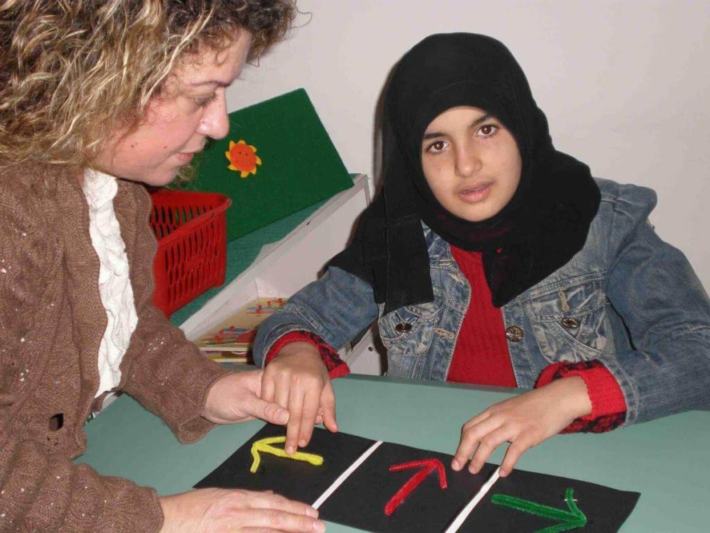 A girl with a head scarf examines a tactile board with 3 arrows on it with her teacher.