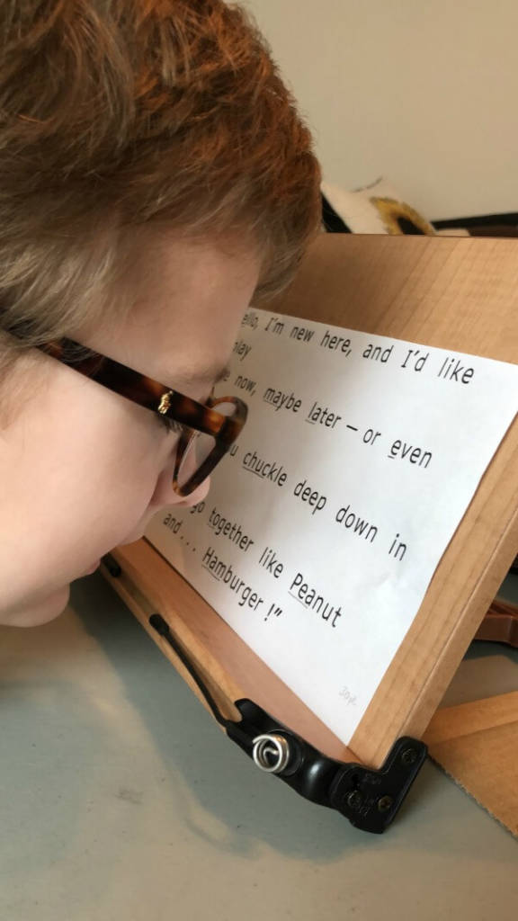 A boy with glasses reads large print text on a slant board at a distance of 2-3 inches.
