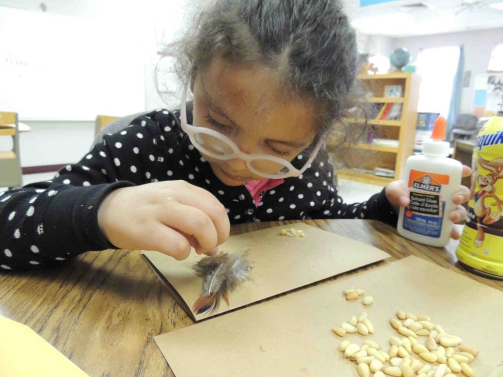 A young girl with glasses examines a feather while holding a bottle of glue.