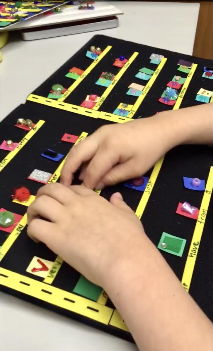 Aidan reads braille whole-word contractions written with textures. A student’s hands track across rows of textures displayed on a velcro board.