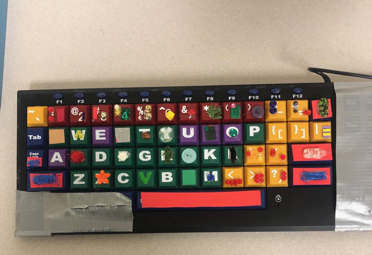 A computer keyboard has small textures velcroed to the keys. Other parts of the keyboard are covered with silver duct tape.
