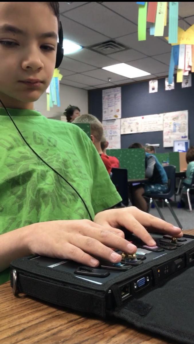 Aidan uses a BrailleNote to take a spelling test in his general education class in 2018. A student wearing headphones uses a BrailleNote adapted with textures on each key.