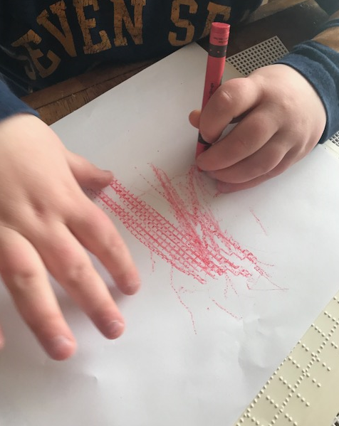 Liam rubs a red crayon on brailled grid paper