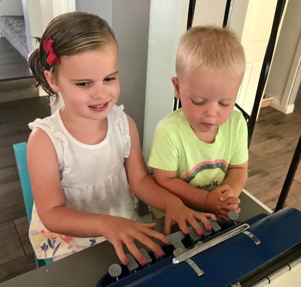 A young girl uses a Perkins braillewriter while her younger brother looks on.