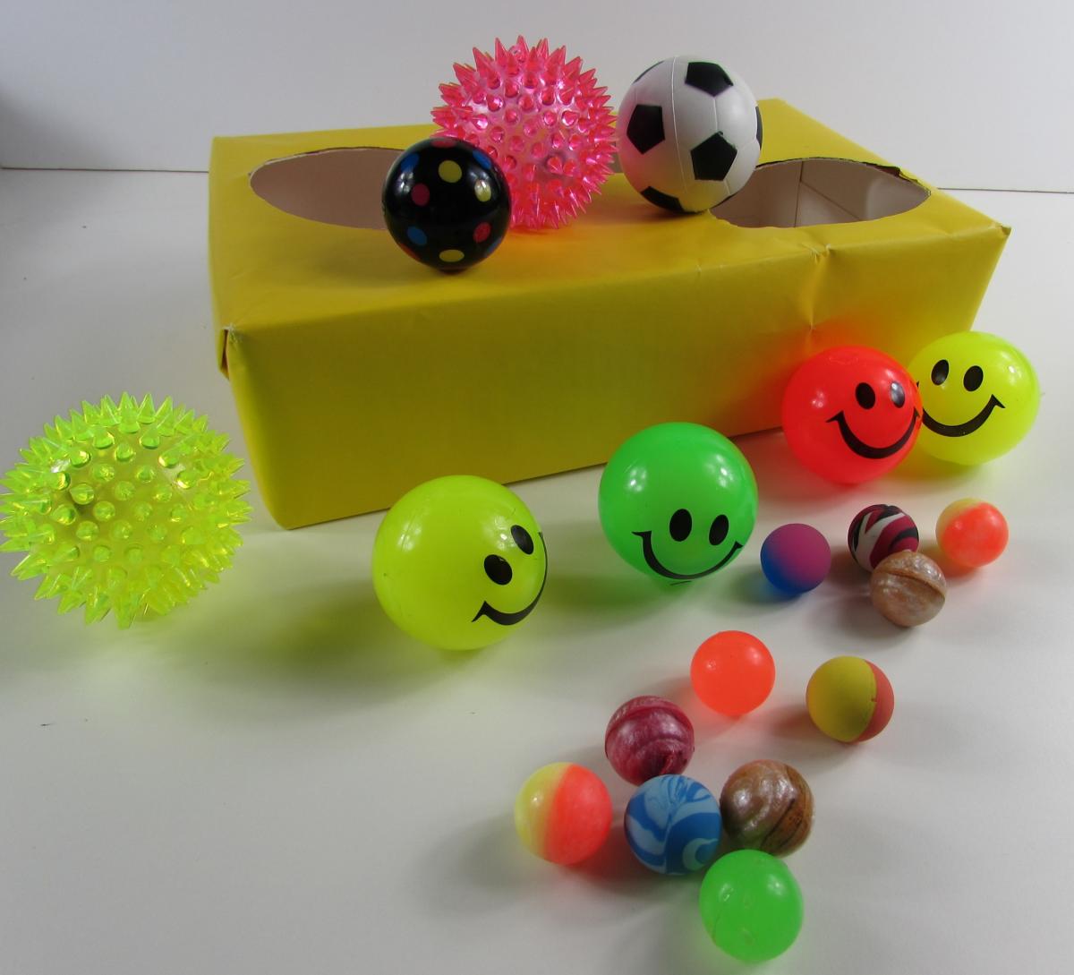 Homemade shape sorter from a cardboard box covered in yellow wrapping paper with three holes cut in the top of the box, Big, Medium and Little.  A selection of bright coloured balls are on and around the box 