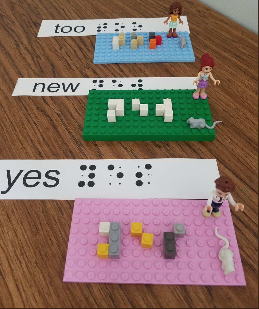 Sight words “too” “new” and “yes” are spelled out on Lego bases with single, double, and triple Lego pieces.  Flash cards with large print, braille cells, and embossedbBraille are shown next to the base.  Lego girl characters and their pet mice are placed on the board as well.