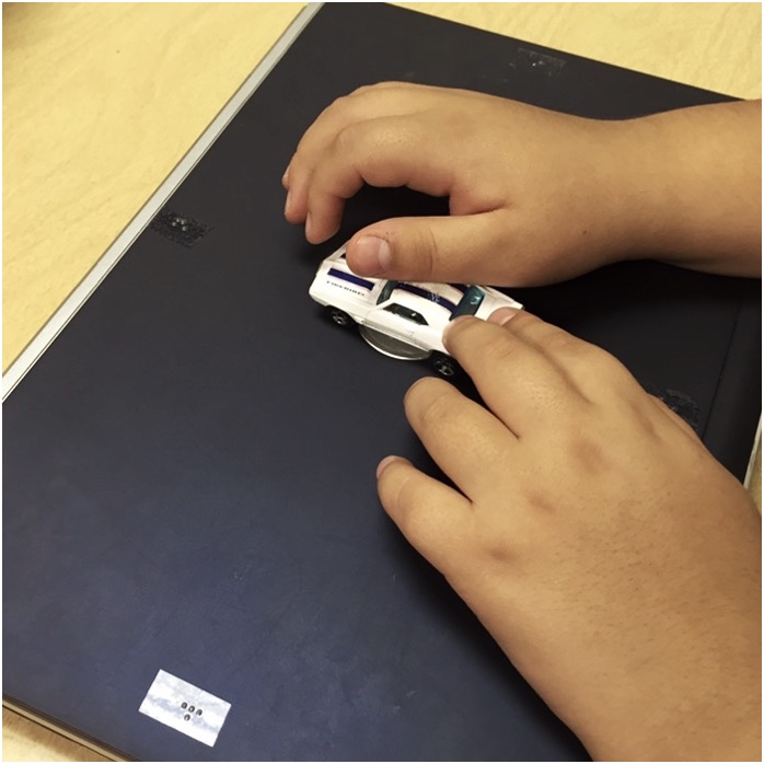 student's hands push a toy car north on top of a closed laptop with directions labeled in braille