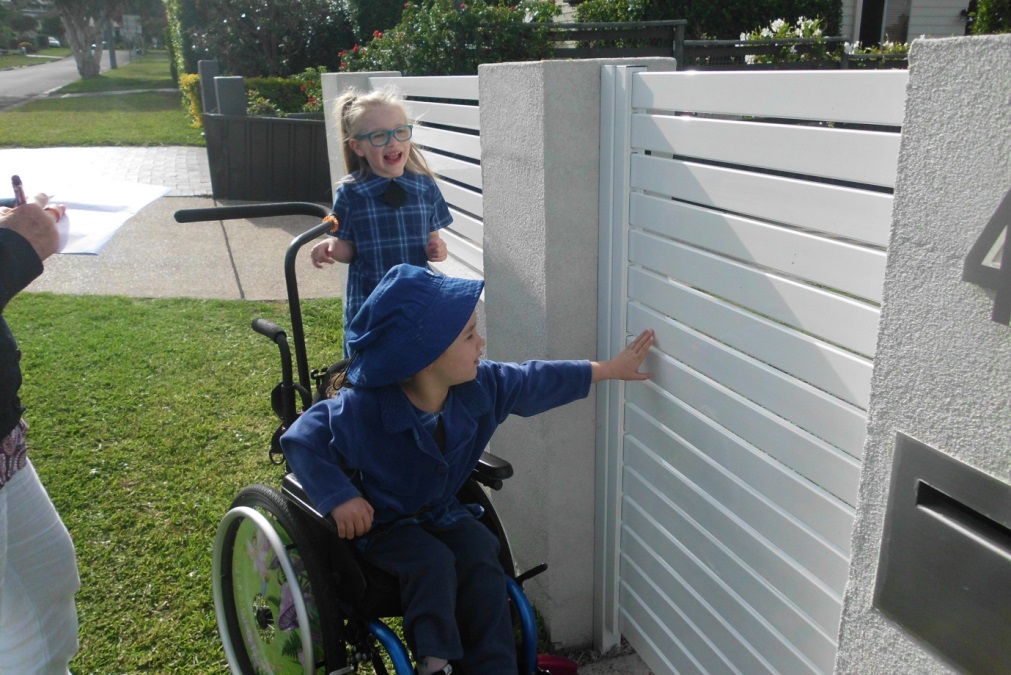 Sarah and Chloe at a fence. Two young girls, one in a wheelchair and the other standing next to the wheelchair.
