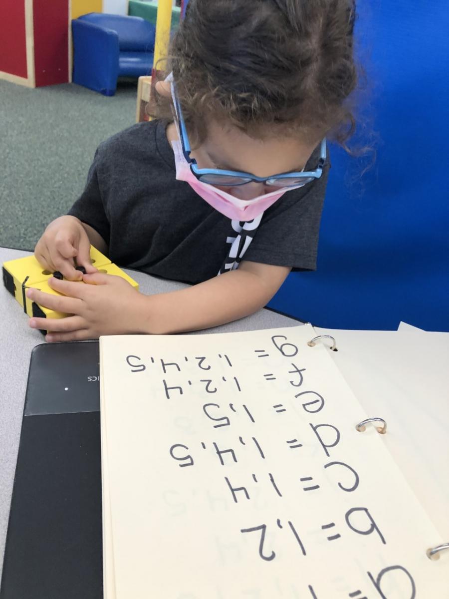 A girl places pegs in a swing cell while looking at a book with print letters of the alphabet and corresponding braille dots.