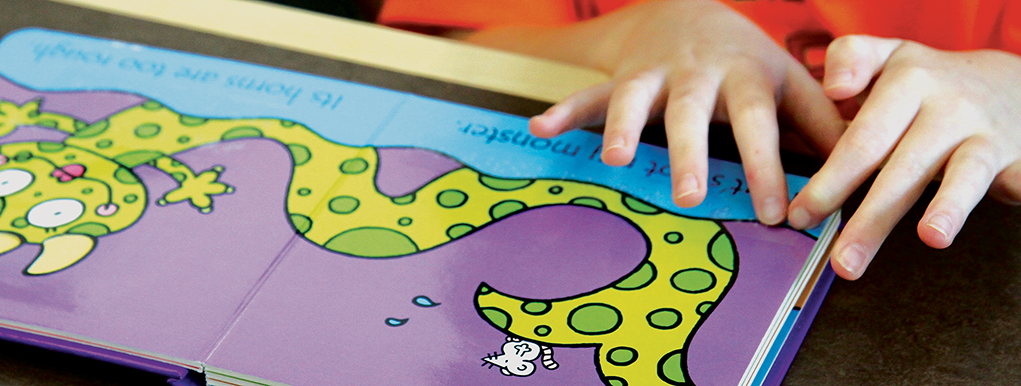 Picture of a board book with two small hands locating the beginning of a line of braille.