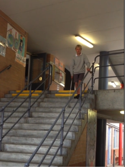 A photo shows a teenage boy in school uniform at the top of a set of stairs. He is descending. He holds the rail in his left hand and his long cane in his right hand.