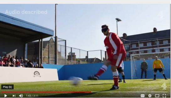 A screen shot from “We are the Superhumans” (see link below) shows a Blind Soccer player kicking a ball while behind him a goalie stands poised. In the grandstand people watch the game. In the top left print reads “Audio described”.