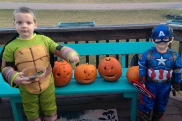 Two brothers in their Halloween costumes next to Jack-o-Lanterns on a bench.