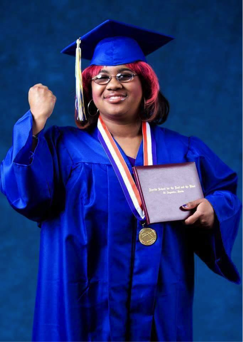 A young woman in a cap and gown holds up her diploma.