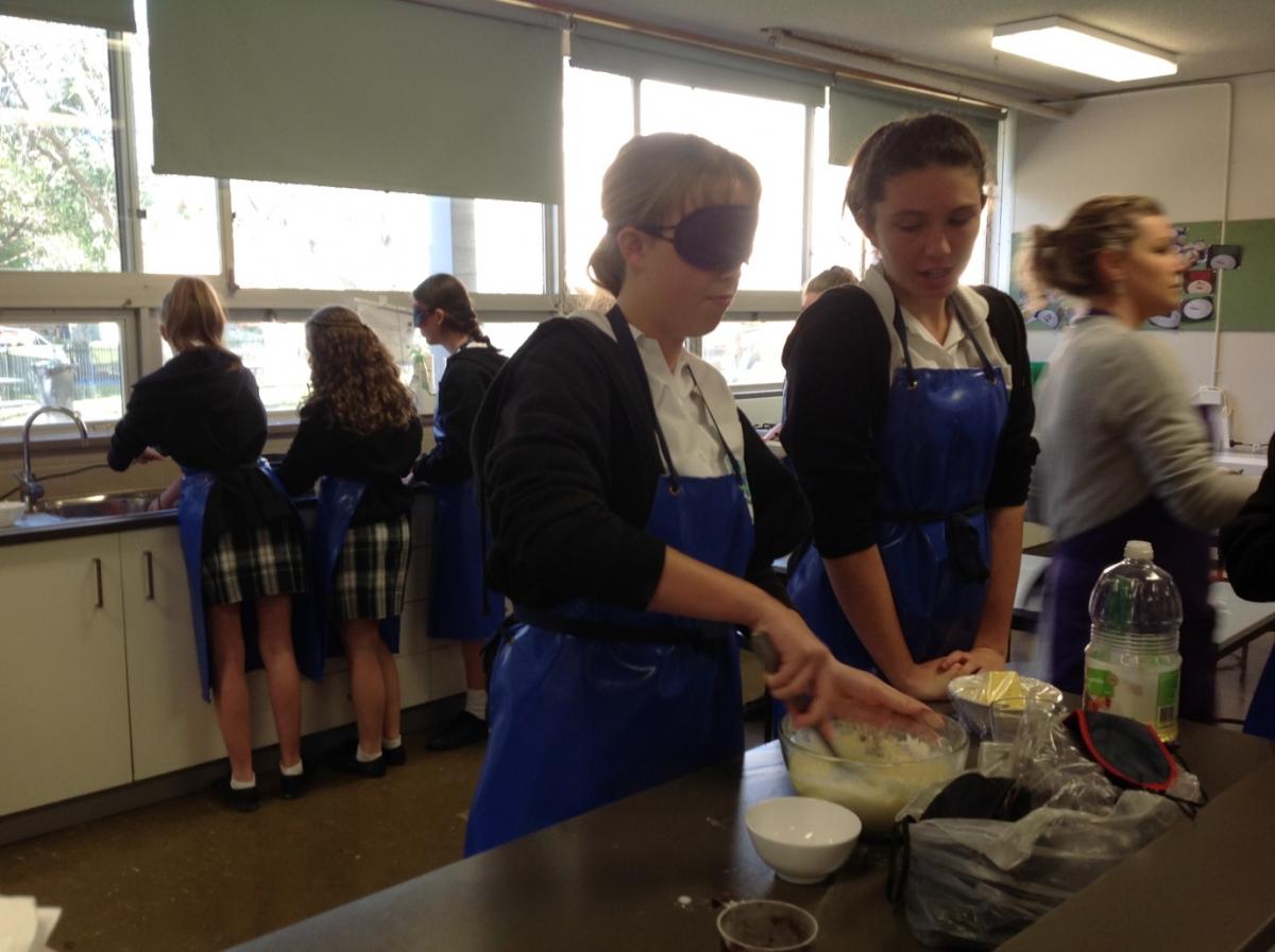 Two students prepare pikelet batter; one (under blindfold) mixes ingredients in a glass bowl while her partner looks on. two students, one is blindfolded, mix the ingredients for their recipe