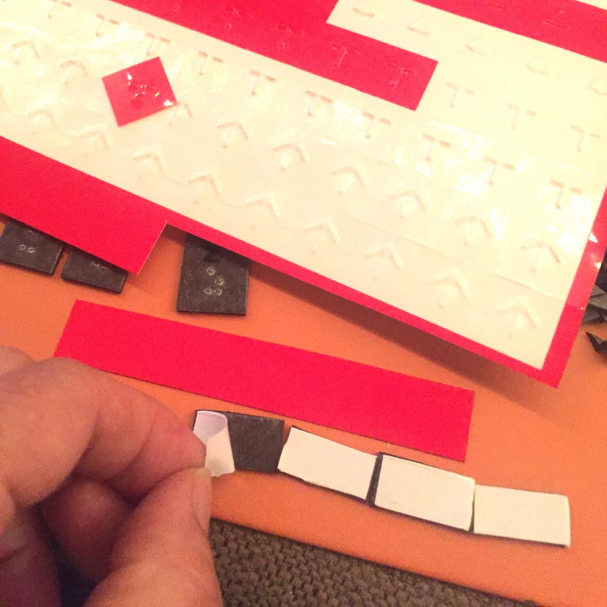 four rectangles lined up horizontally, long red rectangle ready to stick on top, I'm starting to peel paper off magnet to reveal adhesive