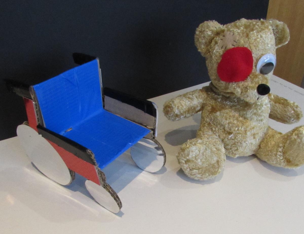 teddy bear with eye patch sitting next to cardboard wheel chair
