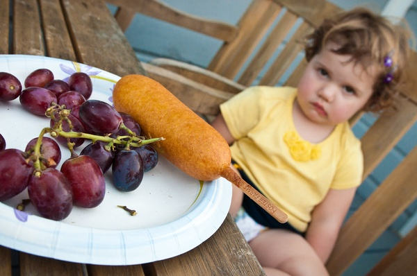 A young girl pouting in the background with a plate of grapes and a corndog in the foreground.