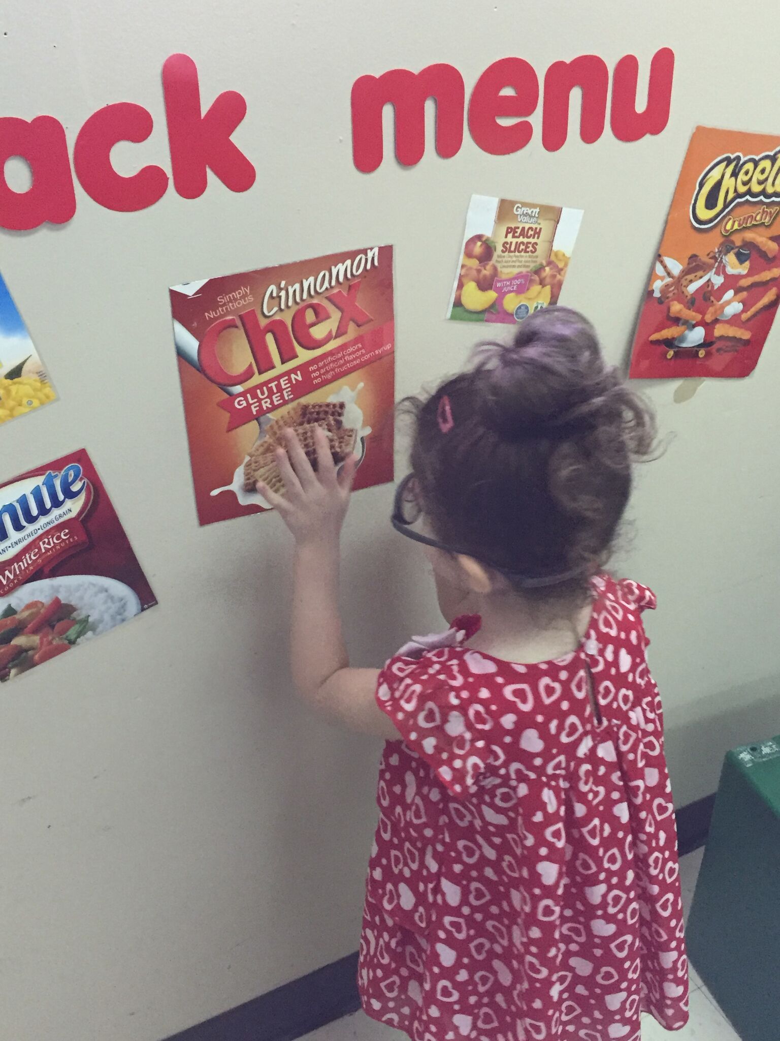 A girl chooses Cinnamon Chex from the Snack Menu wall.