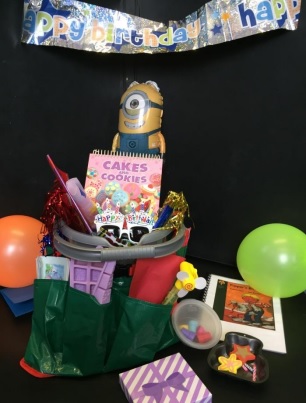 a bucket with supplies for a birthday party including a banner, a book, balloons, and a present