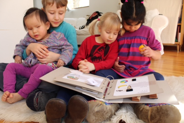 four children looking at the tactile book, they are sitting on the floor and the youngest is sitting in one of the older children's lap