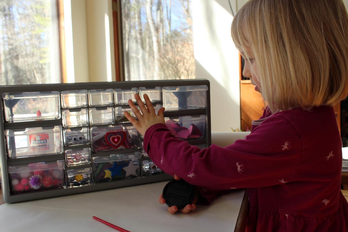 a young girl is touching a plastic set of drawers on top of the table. the drawers are full of arts and crafts supplies.