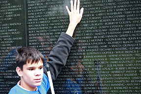 a teen boy holding a white cane strokes the surface of the Vietnam Veterans Memorial