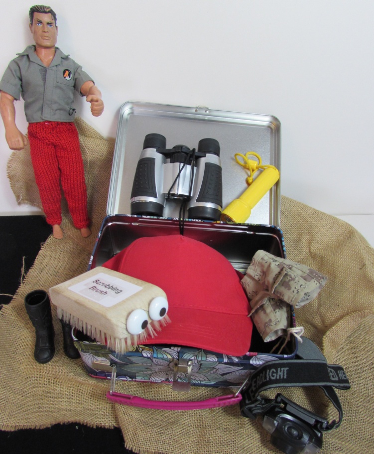 Traction Man with his open equipment tin containing binoculars, torch, head torch, hat, boots, shorts, and string