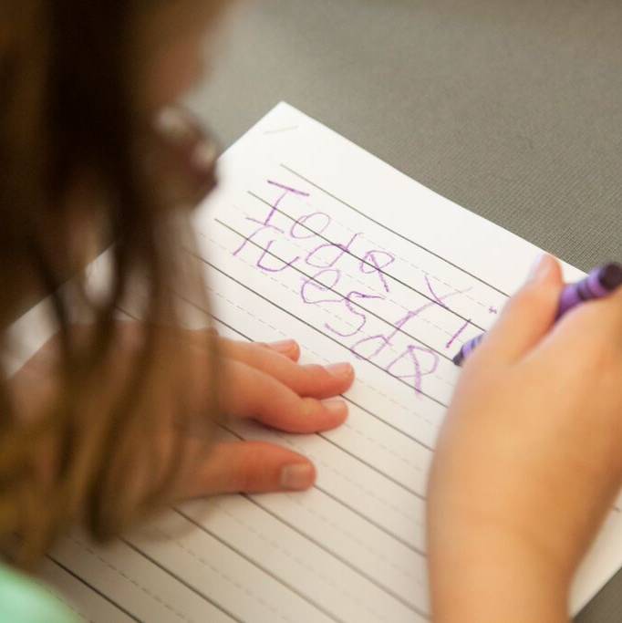 A girl writes the word “Tuesday” with a purple crayon on lined paper.