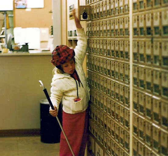 Anna with cane in one hand reaches up with her other hand to get the mail out of a post office box.
