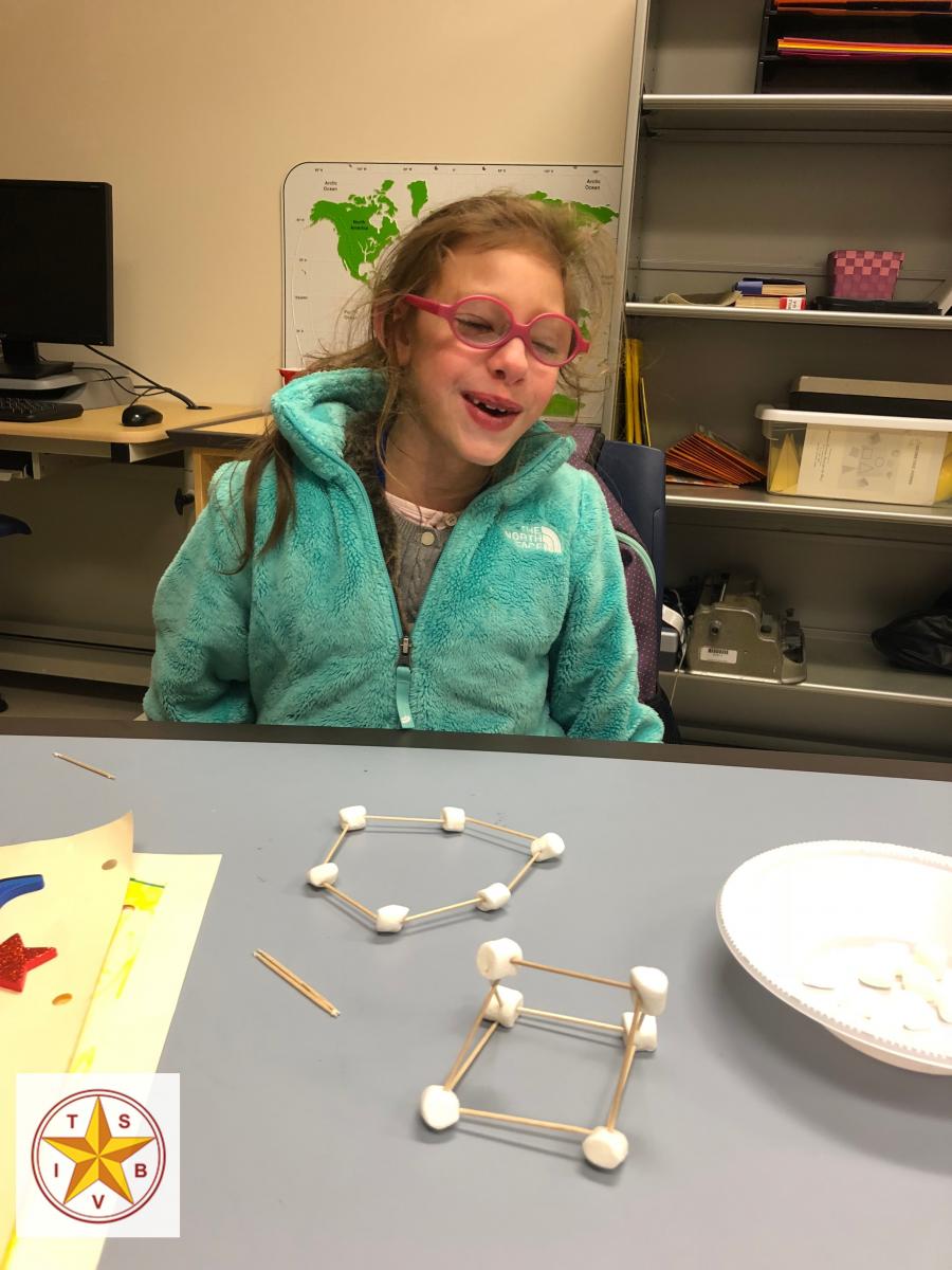 A girl with glasses at a work table with shapes made of marshmallows and sticks
