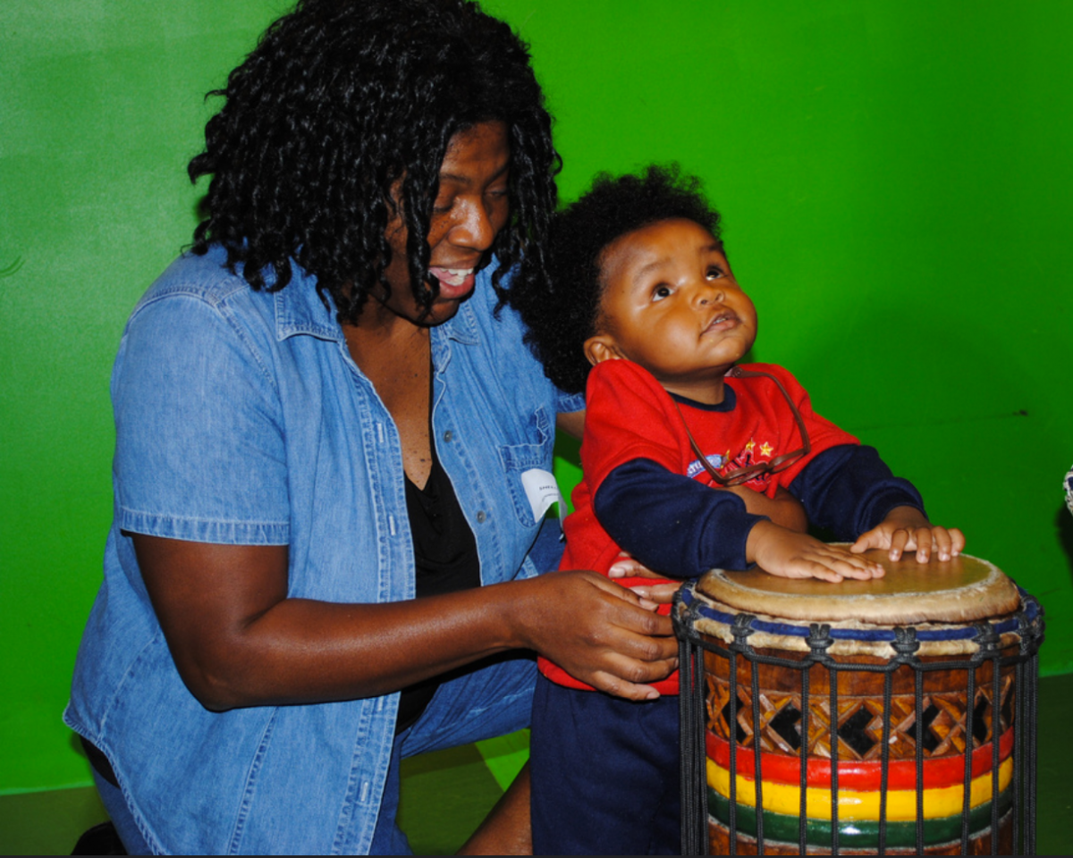 A mother kneels next to her young son playing the drums
