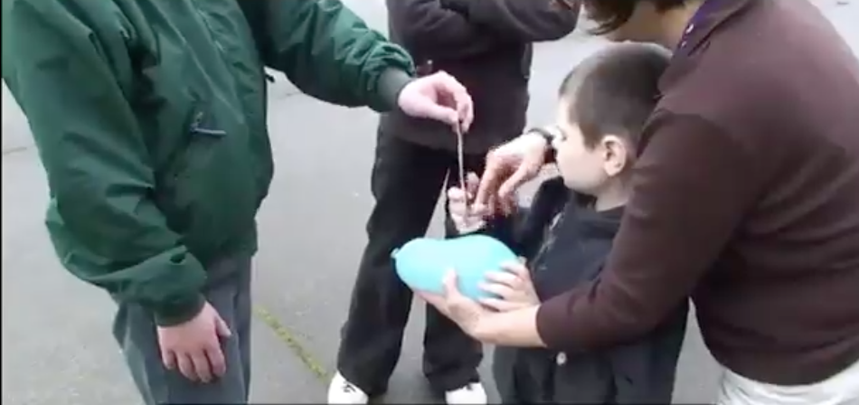 A boy holds a water balloon with one hand while reaching for a fork with the other hand.