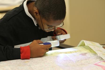 Teenage boy with glasses uses two handheld magnifiers to look at a map.