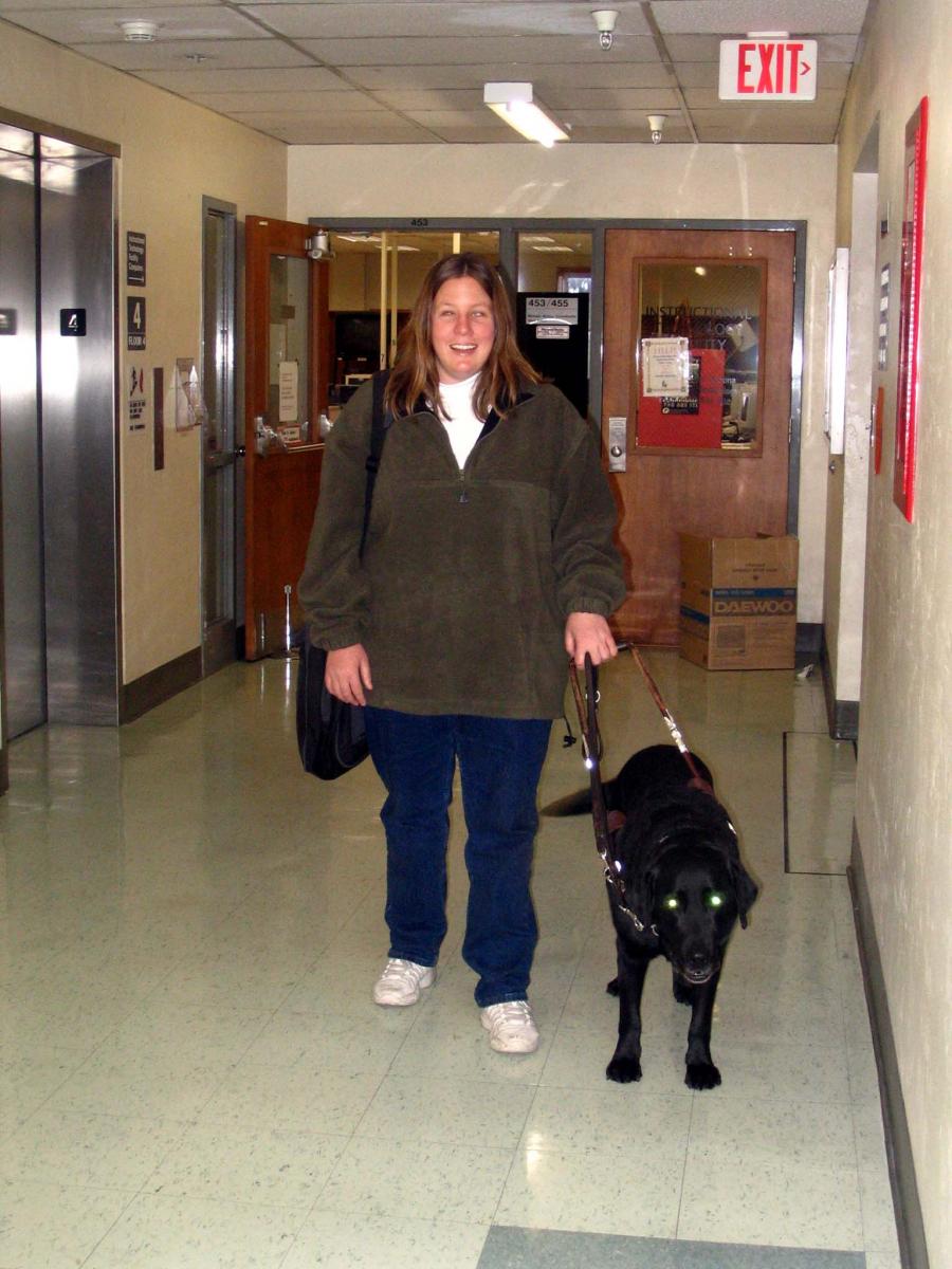 Anna and her dog guide walk down a hallway in a college  building.