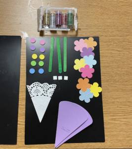 Crafting supplies for a flower craft that includes black paper for the background, lace, colorful flowers, texture stickers, glitter, and popsicle sticks for the stems