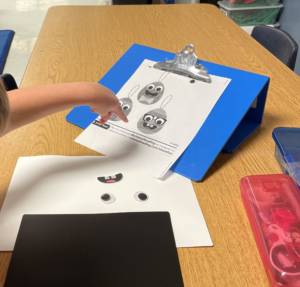 Student at table pointing to instructions on a slant board to picking googly craft eyes and a mouth sticker to complete craft.
