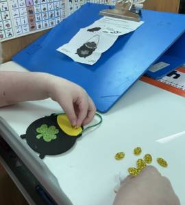 Student making a foam "pot of gold" craft with a shamrock on the black pot and adding foam gold coins. Student has a slant board with written direction on how to complete the craft.