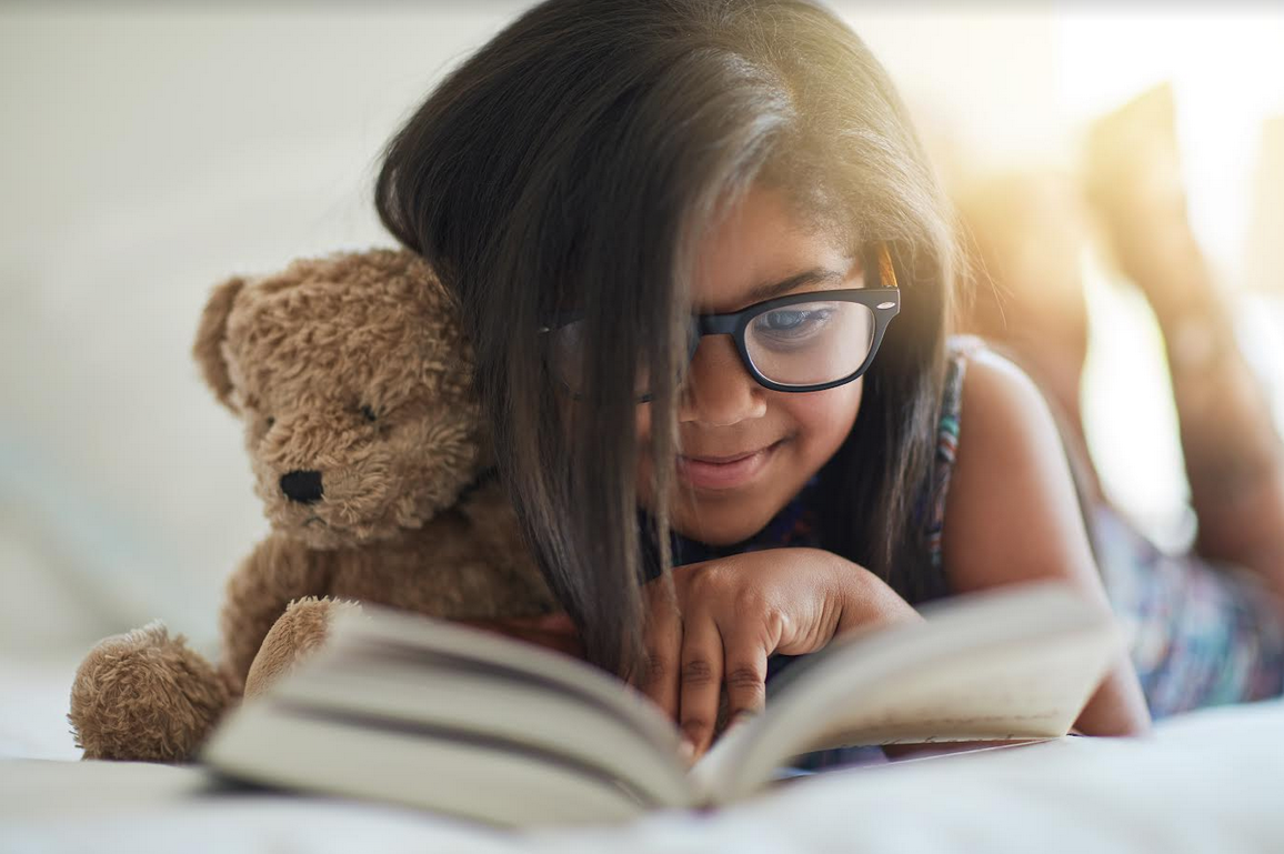 A little girl on happily reading a book holding a teddy bear