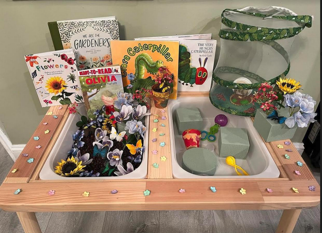Butterfly and flowers sensory bin with plastic flowers and butterflies together with coffee beans in a two bin table. The other bin has small pots, scoops and small boxes. Around it are books with the theme and a caterpillar/butterfly house.