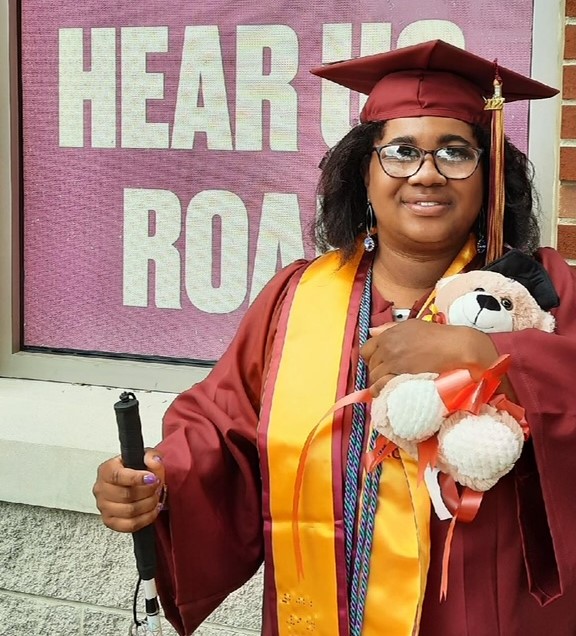 Jasmyn in her cap and gown, with her cane and holding at teddy bear, at Kutztown University