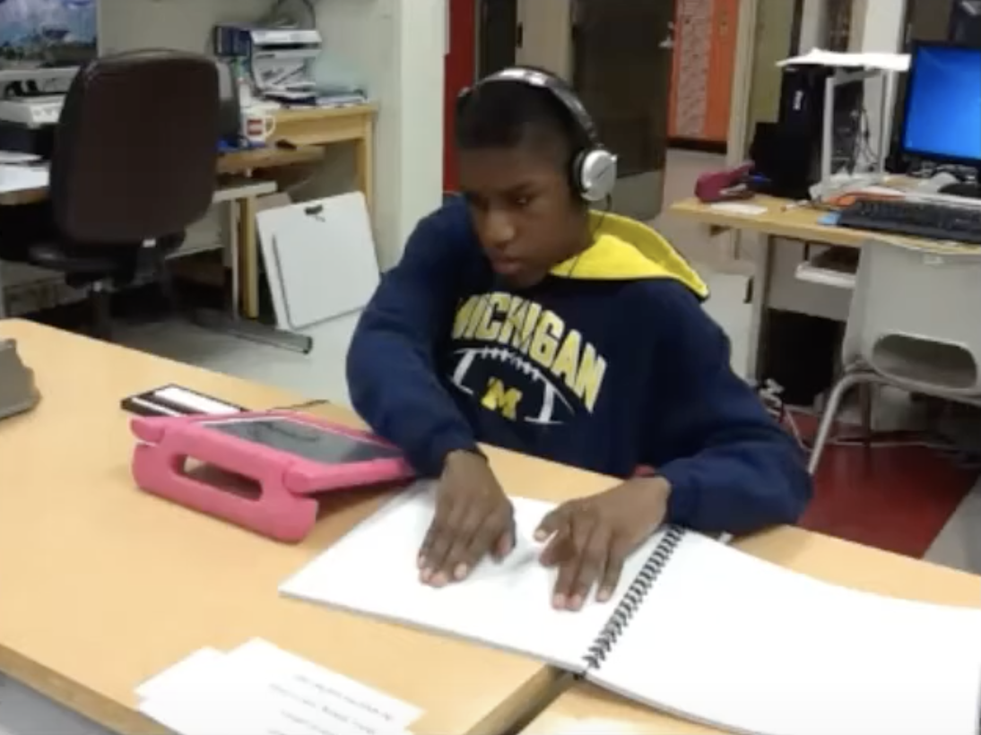 A boy wearing headphones reads a braille text next to a tablet.