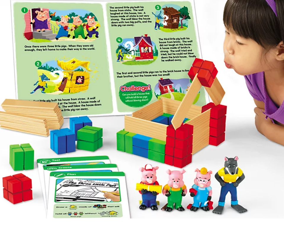 Laskeshore Three Little Pigs STEM kit that includes wooden building type blocks, plastic wolf and three pigs and instruction cards. The picture includes a little girl trying to blow down the house she made. 