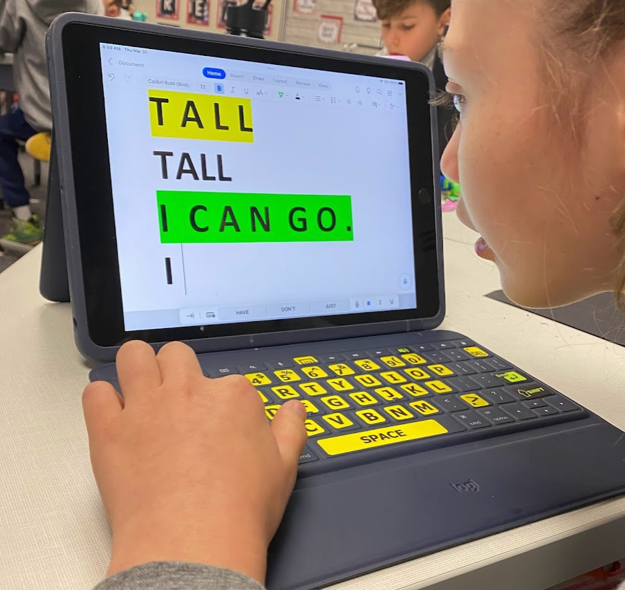 Girl typing on a keyboard with an iPad. Keyboard has large print stickers on it. The screen has large font and highlighted words: Tall and I can go.