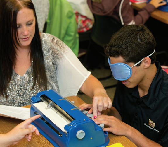 Dr. Nicole Johnson-Mest teaching a student who has a blindfold over their eyes to type on a braille machine. 