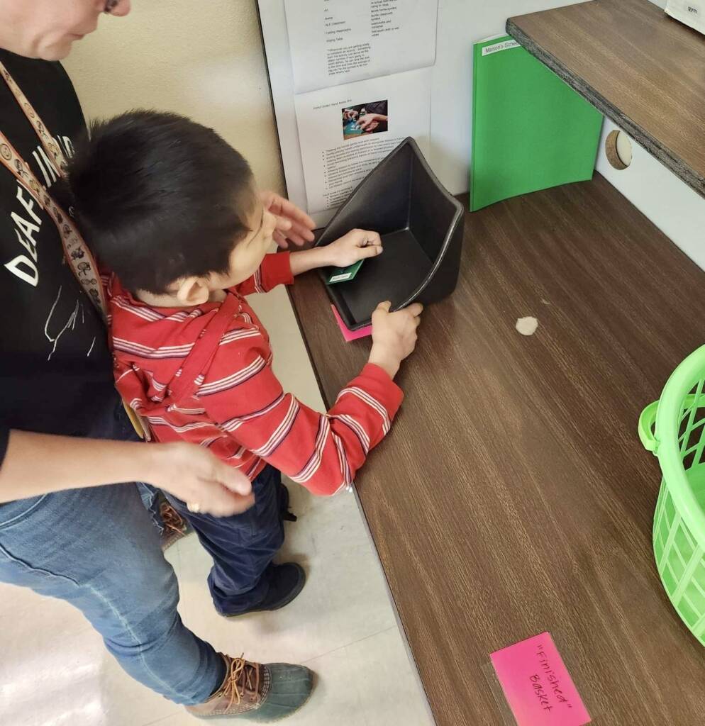 Student standing at table assisted and touching a communication card.