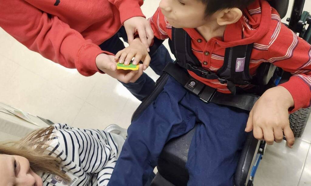 Student in wheelchair touching a piece of the communication system with the help of his teacher. 
