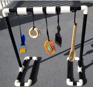 Black PVC frame with white connectors and 5 itmes hanging from the top on black elastic; a roller, 3 wooden rings, a bright orange sensory toy, a black plastic hair pick, and a slotted wooden spoon. 